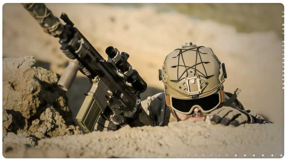A imagem mostra um soldado em um esconderijo, em ambiente de guerra, com uma arma de cano longo na mão. Ele está de capacete e óculos escuro tático. A mão dele com luva é utilizada para apoiar e espiar o horizonte.,